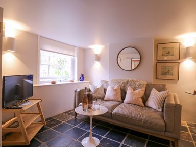 A living room with a gray sofa and pillows a small round table with bottles and glasses a television on a wooden stand and framed pictures on the wall at Mariners Cottage King Street in Aldeburgh
