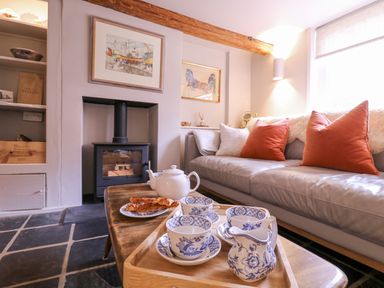 A living room with a sofa and orange cushions a wooden coffee table with tea set and pastries a fireplace and framed pictures on the wall at Mariners Cottage King Street in Aldeburgh