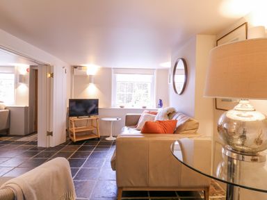A living room with a beige sofa and orange pillows a glass table with a lamp and a TV on a wooden stand at Mariners Cottage King Street Aldeburgh