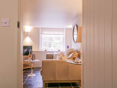View into a living room with a beige sofa with cushions a round side table with wine bottle and glasses a glass top table with a lamp and mirror on the wall at Mariners Cottage King Street in Aldeburgh