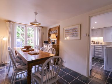 A dining room with a wooden table and white chairs next to a cabinet with plates and a lamp at Mariners Cottage King Street Aldeburgh