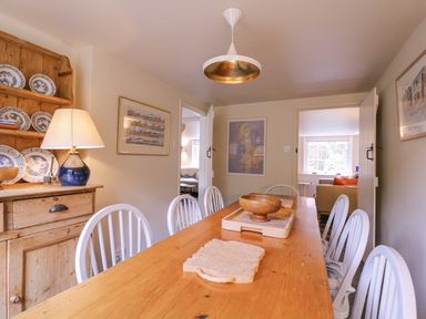 A dining room with a wooden table and white chairs a cupboard with blue and white plates and a lamp on it at Mariners Cottage King Street Aldeburgh