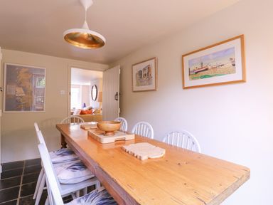 A dining room with a wooden table and white chairs with cushions pictures on the wall and a light fixture at Mariners Cottage King Street Aldeburgh