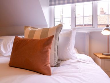 A bed with three pillows next to a window and a bedside table lamp at Mariners Cottage King Street in Aldeburgh