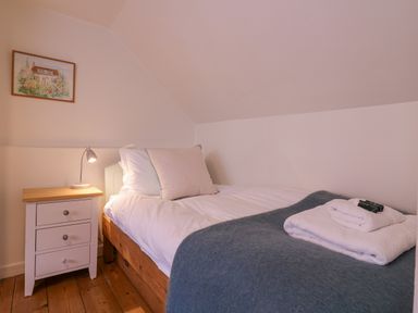 A bedroom with a single bed white linens and a blue blanket next to a wooden nightstand with a lamp and a framed picture on the wall at Mariners Cottage King Street Aldeburgh