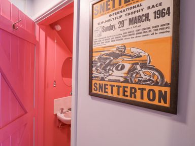 An interior view showing a framed motorcycle race poster on a wall beside a doorway leading to a small room with a pink door and a sink inside