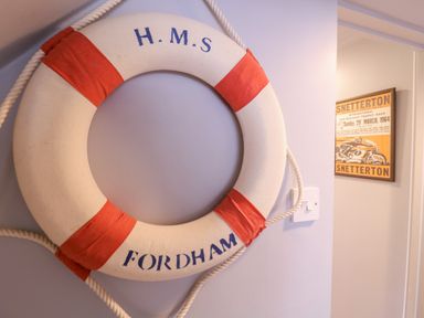 A lifebuoy with red straps and the text H M S Fordham on a wall near a light switch and a framed poster at Mariners Cottage King Street in Aldeburgh