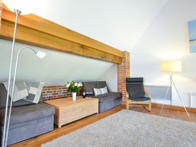 A living room with a gray sofa a wicker coffee table with flowers and wine a black armchair and lamps at Two Chantry Barns Orford
