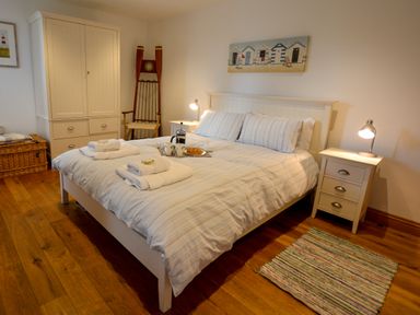 A bedroom with a double bed and striped bedding two bedside tables with lamps a wicker basket a wooden chair a white wardrobe and a small rug on wooden floor at Two Chantry Barns Orford