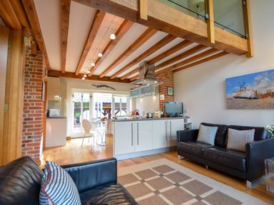 An open living room and kitchen with black leather sofas wooden beams and a painting of a boat at Two Chantry Barns Orford