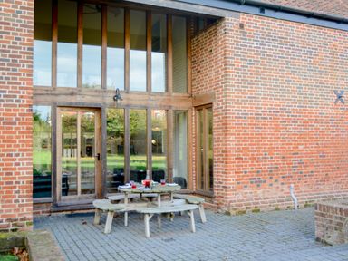 An outdoor patio area with a round wooden picnic table and benches in front of a large glass door and brick wall at Two Chantry Barns Orford