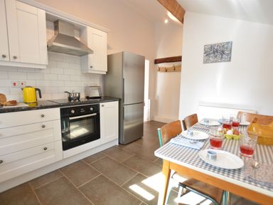 A kitchen with white cabinets a stainless steel refrigerator and an oven next to a dining table with plates glasses and chairs at Wheelwright's Barn in Walpole Near Halesworth