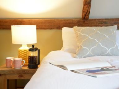 A bedside table with a lamp two mugs and a coffee press next to a bed with pillows and an open book at Wheelwright's Barn in Walpole Near Halesworth