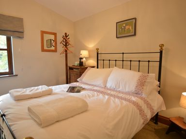 A bedroom with a double bed with white linens and folded towels a wooden bedside table with a lamp a coat rack and a window at Wheelwrights Barn Walpole Near Halesworth