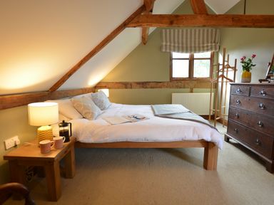 A bedroom with a wooden bed white linens two pillows a wooden nightstand with a lamp two mugs and a coffee press a wooden chest of drawers and a window with a striped blind at Wheelwright's Barn Walpole Near Halesworth