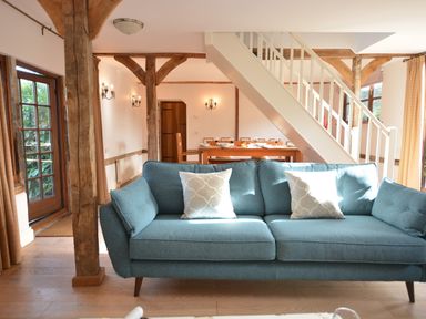 A living room with a blue sofa and white pillows near wooden beams and a dining table under stairs at Wheelwright's Barn in Walpole Near Halesworth