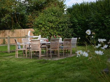 An outdoor dining table with eight chairs on grass surrounded by trees and a wooden fence at Wheelwright's Barn in Walpole Near Halesworth