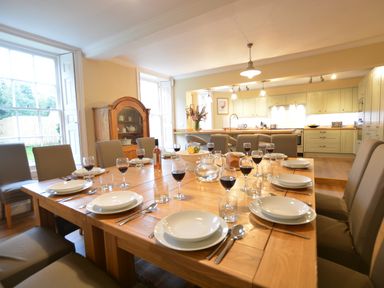 A dining room with a large wooden table set with plates glasses and cutlery and a kitchen with beige cabinets in the background at Henstead Hall in Henstead near Beccles
