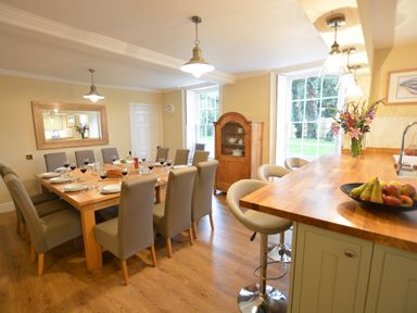 A dining room with a wooden table set for a meal and a kitchen island with bar stools and fruit bowl at Henstead Hall Henstead near Beccles