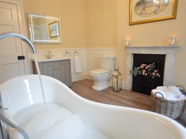 A bathroom with a bathtub filling with water a toilet a sink with mirror a basket with towels and a fireplace with flowers at Henstead Hall in Henstead near Beccles