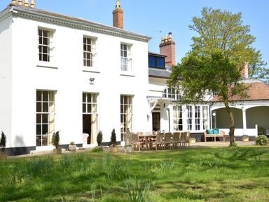 The exterior of a white two-story house with large windows a patio with outdoor wooden furniture and a tree in the garden at Henstead Hall in Henstead near Beccles
