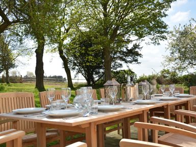 An outdoor dining table set with plates glasses cutlery and decorative lanterns surrounded by wooden chairs under trees at Henstead Hall in Henstead near Beccles