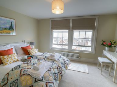 A bedroom with a bed with folded towels pillows and a tray a window with blinds a lamp a painting on the wall and a dressing table with stool and flowers at 10 Church Walk Aldeburgh