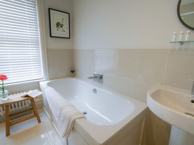 A bathroom with a bathtub, a white towel on the bathtub, a wooden stool with a flower vase and a book, a sink, and a window with blinds at 10 Church Walk, Aldeburgh in Aldeburgh