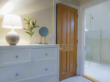 A chest of drawers with a lamp a flower vase and a small round mirror next to a wooden door and an adjoining bathroom with a glass shower enclosure at 10 Church Walk Aldeburgh