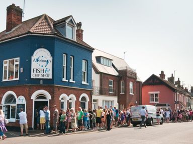 A long line of people outside a blue and brick fish and chip shop on a street with other brick and pink buildings at 10 Church Walk Aldeburgh