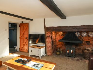 A living room with a wood burning stove in a brick fireplace a wooden door a television on a white cabinet and a wooden table at 2 Greensleeves in Benhall Near Friston