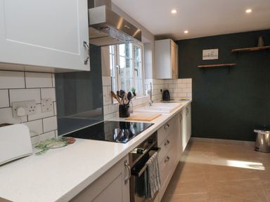 A kitchen with white cabinets utensils on the counter and two wooden shelves on a dark wall at 2 Greensleeves in Benhall Near Friston