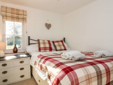 A bedroom with a bed covered in a plaid blanket and pillows with a nightstand and window at 2 Greensleeves in Benhall Near Friston