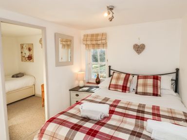 A bedroom with a double bed with plaid bedding and rolled towels on it a bedside table with a lamp and a window showing outdoors plus a doorway to another room with a single bed at 2 Greensleeves in Benhall Near Friston