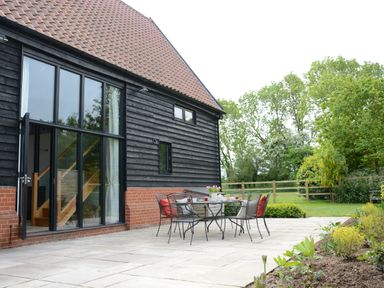 An outdoor patio with a table and chairs next to a black wooden house with large windows and a pitched roof at Manor House Barn Peasenhall