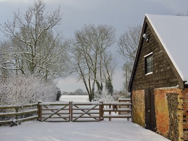 A snow-covered wooden gate and fence next to a brick and wooden building with a snowy roof and leafless trees in the background at Manor House Barn Peasenhall