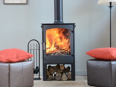 A room with a wood burning stove with fire inside two brown ottomans with red cushions and a lamp in Manor House Barn Peasenhall