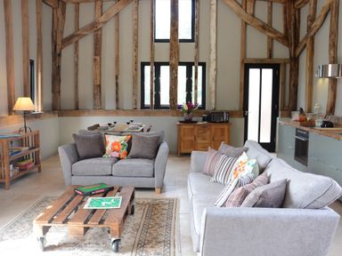 A living room with two gray sofas a wooden coffee table with a scrabble game a dining table set with plates and wine glasses a wooden cabinet with a microwave and flowers kitchen area with stove and utensils at Manor House Barn Peasenhall