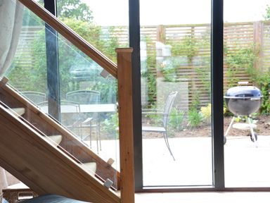 A sliding glass door opening to a patio with metal chairs a table and a barbecue grill behind a wooden staircase with glass panels at Manor House Barn Peasenhall