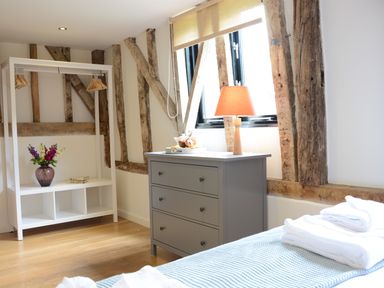 A bedroom with a grey dresser with a lamp on it and a white open wardrobe with hangers and flowers at Manor House Barn Peasenhall