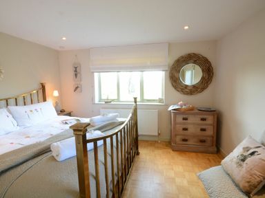 A bedroom with a brass bed, wooden chest of drawers, window with white blind, and a bench with a bunny pillow at The Nookery at Snape Hall in Snape Suffolk