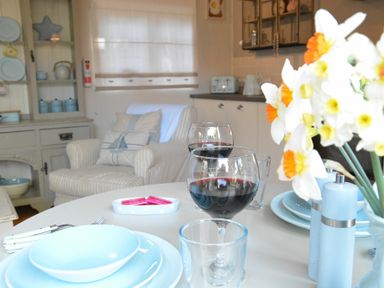 A dining table with plates wine glasses and flowers in a room with an armchair and kitchen cabinets at The Nookery at Snape Hall in Snape Suffolk