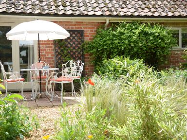A garden patio with white metal chairs and a table under a white umbrella in front of a brick building with greenery at The Nookery at Snape Hall in Snape Suffolk