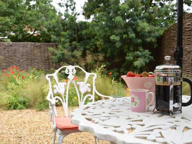 A white metal garden table with a coffee press a pink cup and a bowl of strawberries next to a white metal chair with a cushion in a garden at The Nookery at Snape Hall in Snape Suffolk