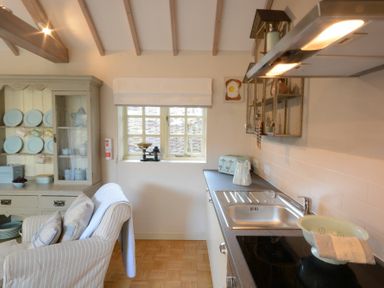 A kitchen with a sink stove and cupboard next to a striped armchair and a hutch with plates at The Nookery at Snape Hall in Snape Suffolk