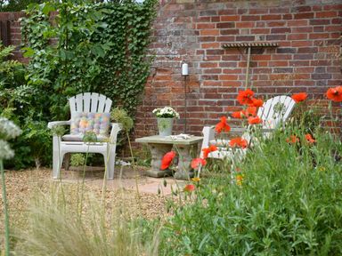 A garden with two white chairs a small stone table and red flowers by a brick wall at The Nookery at Snape Hall in Snape Suffolk