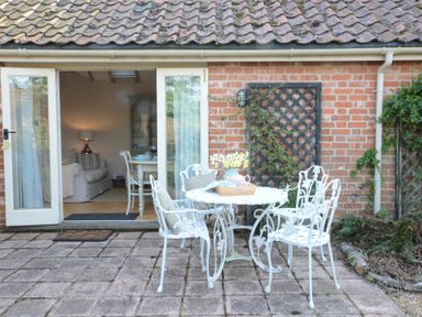 An outdoor patio with a white metal table and four chairs in front of glass doors leading to a dining room at The Nookery at Snape Hall in Snape Suffolk