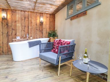 A wooden deck area with a white bathtub a grey cushioned chair with red pillows and a round table with a bottle and two wine glasses at Yew Tree Farm Stable Worlingworth near Framlingham