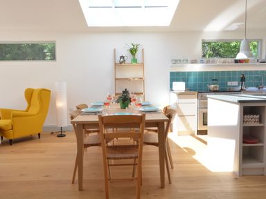 A dining area with a wooden table set with plates and glasses next to a kitchen with teal tiles and a yellow armchair in 2 Pink Cottages Aldeburgh