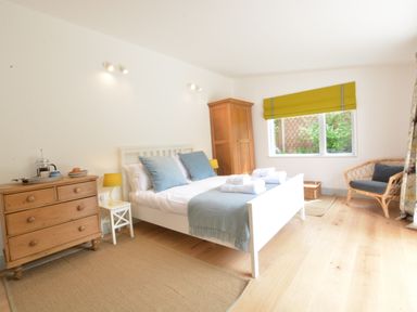 A bedroom with a white bed and blue pillows next to a wooden dresser and a wooden chair at 2 Pink Cottages Aldeburgh